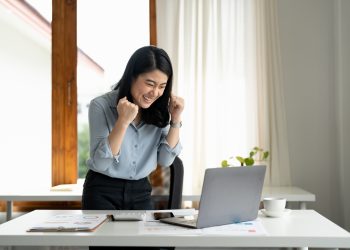 Excited young woman standing at table with laptop and celebrating success. achieving goals excited