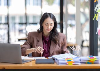 Freelancer Happy business Asian woman in knitwear taking notes at laptop sitting at desk office,