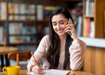 Job hunting. Happy arab woman talking by smartphone with hr representetive and taking notes, sitting