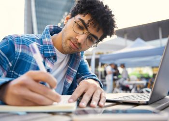 Pensive middle eastern freelancer using laptop computer, taking notes, working online at workplace
