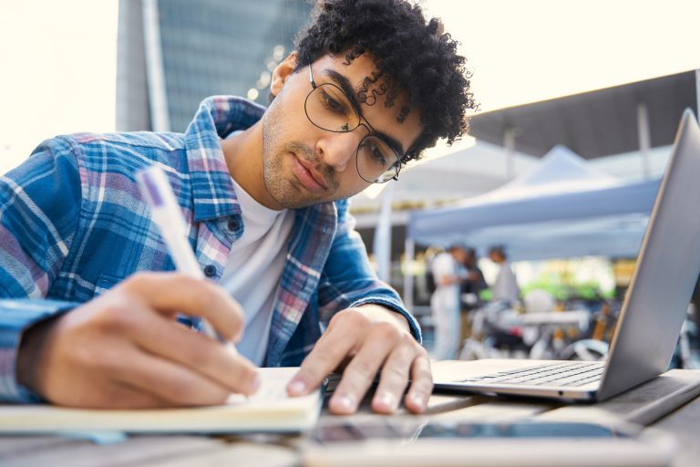 Pensive middle eastern freelancer using laptop computer, taking notes, working online at workplace