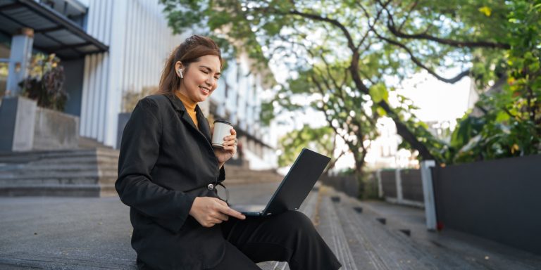 Smiling young Asian business woman using laptop sitting outdoor. hybrid working, searching job