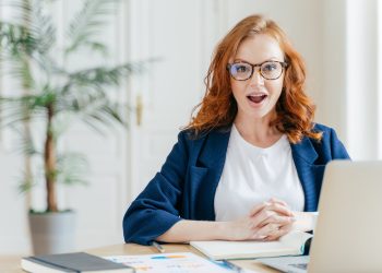Surprised female employee has job, develops new business strategy, poses in front of opened laptop