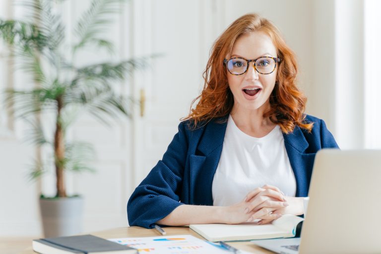 Surprised female employee has job, develops new business strategy, poses in front of opened laptop