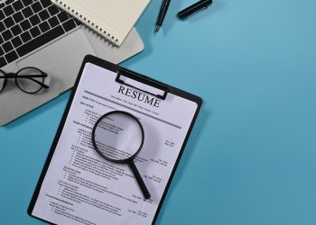 Top view laptop computer, magnifying glass and resumes applicants on blue background.