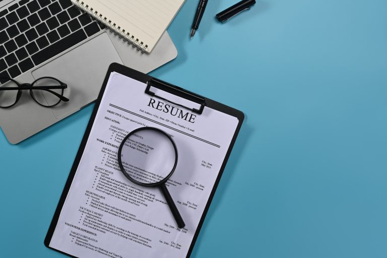 Top view laptop computer, magnifying glass and resumes applicants on blue background.