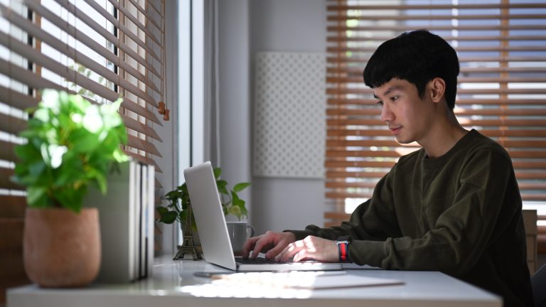 Young man freelancer searching online information, working with laptop computer at home office.