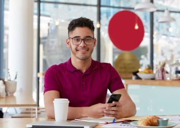 Handsome cheerful young man searches job, browses webpage on modern cell phone, checks information i