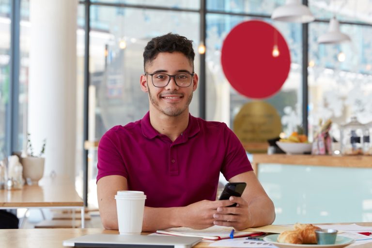 Handsome cheerful young man searches job, browses webpage on modern cell phone, checks information i